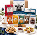 A curated tea and biscuit gift hamper arrangement on a white pedestal, featuring an Evercove Gifts box, a Bridgette Afternoon Tea box, several Walker's tea bags, a jar of Bridges preserves, cookies, scones, jam, and chocolate squares, with a teacup and floral props around it.